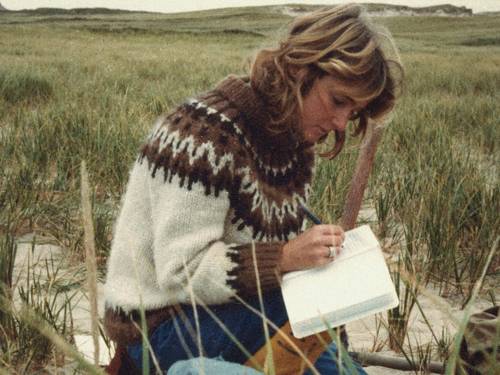 Zu sehen ist eine weiblich gelesene Person, die in einer dünenartigen Landschaft kniet und in ein Notizbuch schreibt.